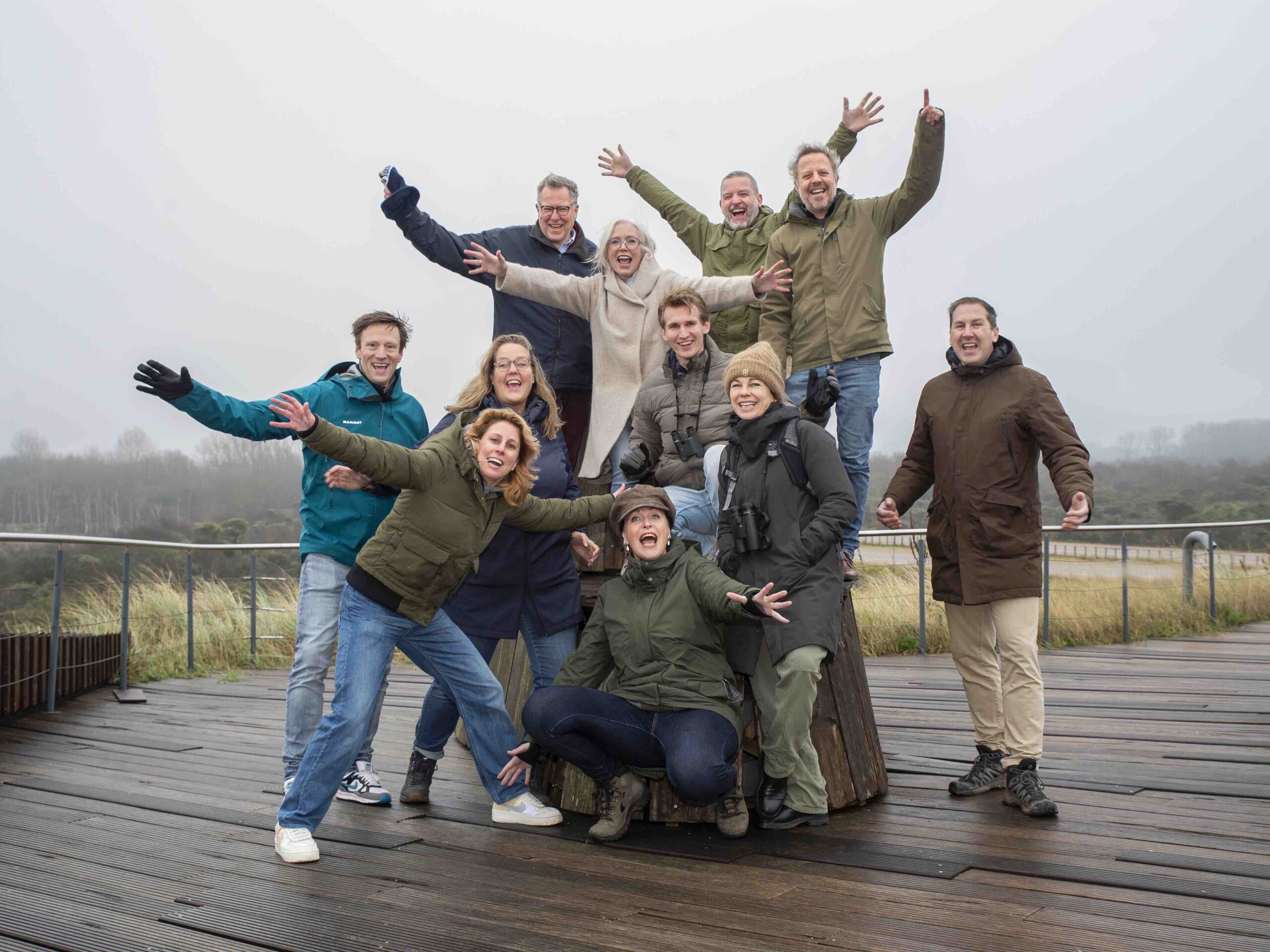 Een groep van elf volwassenen staat buiten op een houten platform en poseert vrolijk voor de camera. Ze dragen warme jassen en winterkleding en steken hun armen omhoog of naar voren in een speelse houding. Op de achtergrond is een mistig, natuurlandschap te zien met gras en lage begroeiing. De sfeer is energiek en gezellig, alsof het om een groepsuitje of gezamenlijke activiteit gaat.