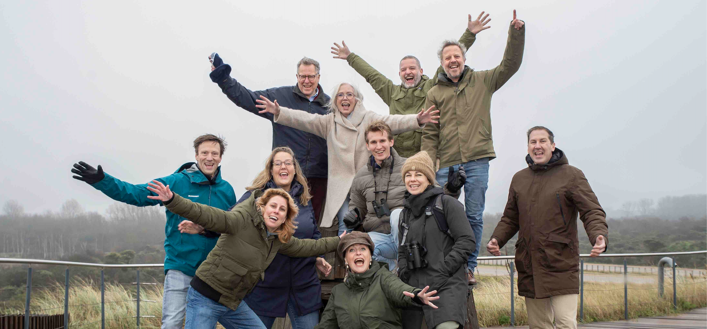 Een groep van elf volwassenen staat buiten op een houten platform en poseert vrolijk voor de camera. Ze dragen warme jassen en winterkleding en steken hun armen omhoog of naar voren in een speelse houding. Op de achtergrond is een mistig, natuurlandschap te zien met gras en lage begroeiing. De sfeer is energiek en gezellig, alsof het om een groepsuitje of gezamenlijke activiteit gaat.